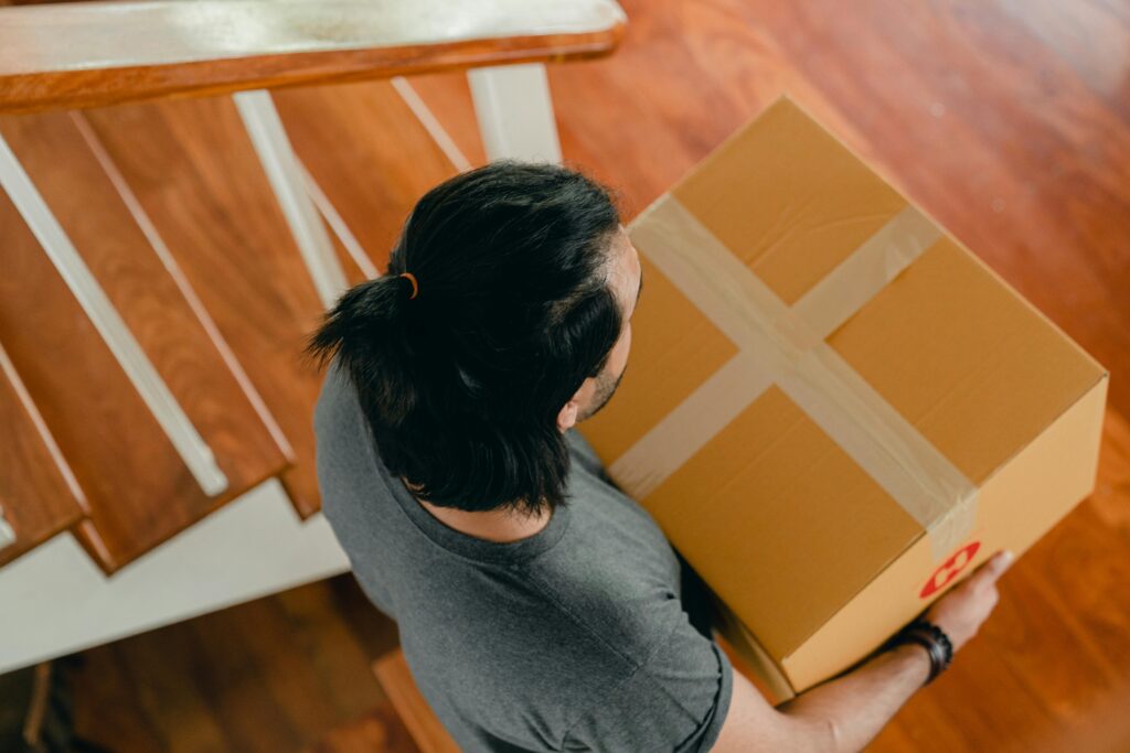 A man with dark hair walks down stairs carrying a cardboard box indoors.
